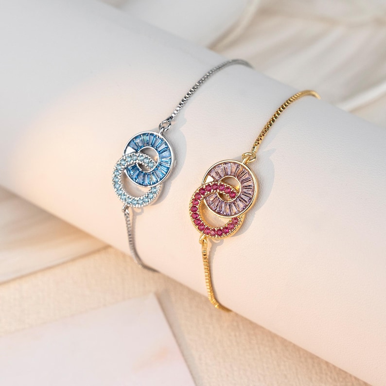 May include: Two bracelets with interlocking circles, one silver with blue stones, the other gold with pink and red stones. The bracelets are displayed on a white surface, showcasing the jewelry's design and color variations.