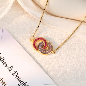 May include: A gold-colored necklace featuring two interlocking circles. One circle is adorned with small, round, red gemstones, while the other is embellished with rectangular, clear gemstones. The necklace is on a white surface with text that reads "Mother & Daughter".