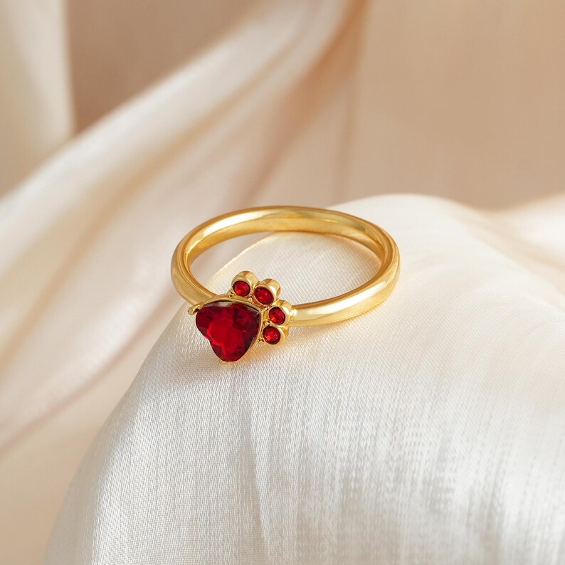 May include: A gold-colored ring featuring a paw print design. The ring has a heart-shaped red gemstone at the center of the paw, with smaller red gemstones forming the paw pads. The ring is set against a white fabric background.