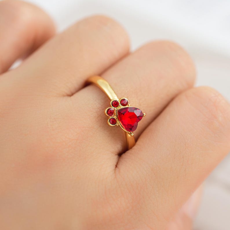 May include: A gold-colored ring featuring a paw print design. The ring's focal point is a heart-shaped, vibrant red gemstone, complemented by smaller red gems. The ring is designed with a smooth, polished band.