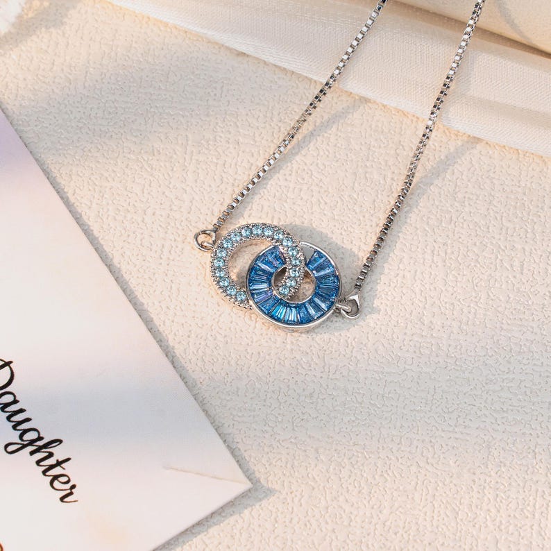 May include: A silver necklace with two interlocking circles, one with light blue crystals and the other with darker blue baguette-cut crystals. The necklace is on a white surface.
