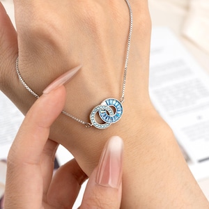 May include: A silver bracelet featuring two interlocking circles, each adorned with sparkling blue crystals. One circle has small, round crystals, while the other has rectangular crystals. The bracelet is displayed on a wrist.