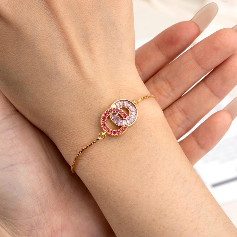 May include: Gold-toned bracelet featuring two interlocking circles, one with pink and the other with clear gemstones. The bracelet has a delicate chain and is designed to be worn on the wrist. A stylish piece of jewelry.