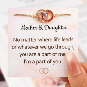 May include: A gold-toned bracelet with two interlocking circles, one with clear and the other with red stones, is displayed on a card. The card reads "Mother & Daughter" and includes a sentimental message. The bracelet is held by a hand.