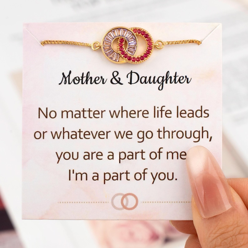 May include: A gold-toned bracelet with two interlocking circles, one with clear and the other with red gemstones, is displayed on a card. The card reads "Mother & Daughter" and includes a sentimental message.