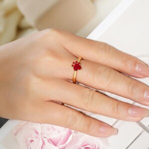 May include: A gold-colored ring featuring a paw print design with red gemstones. The ring is on a finger, showcasing the delicate details of the jewelry. The paw print is a symbol of love for pets.