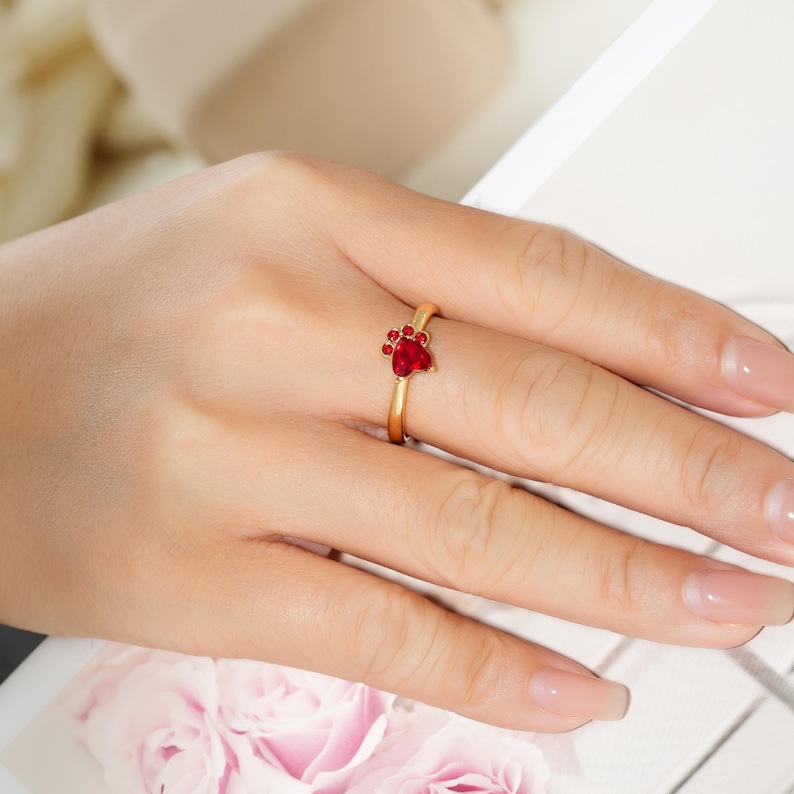May include: A gold-colored ring featuring a paw print design with red gemstones. The ring is on a finger, showcasing the delicate details of the jewelry. The paw print is a symbol of love for pets.