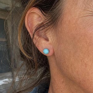Peut inclure: Une petite boucle d'oreille à puce ronde et turquoise vif portée au lobe de l'oreille. La boucle d'oreille présente une surface lisse et polie d'une couleur bleu-vert éclatante, posée sur la peau, avec un design circulaire simple.