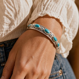 May include: A silver cuff bracelet featuring a series of oval-shaped turquoise stones set in metal frames. The band is decorated with intricate metalwork patterns, including small circular accents between each stone, creating a textured appearance.