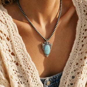 May include: A necklace featuring a string of small, round, dark metallic beads and a central pendant shaped like an arrowhead. The pendant is crafted from silver-toned metal with intricate engravings and holds a smooth, oval, light blue turquoise stone.
