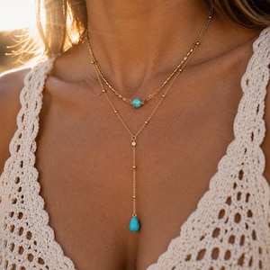 May include: A layered gold-tone necklace featuring a short chain with a round turquoise bead and a longer lariat-style chain with a teardrop-shaped turquoise pendant. The chains are adorned with small gold-tone beads along their length.