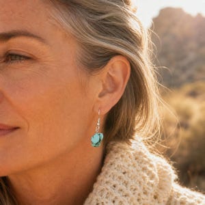 Peut inclure: Une boucle d'oreille pendante composée d'une pierre turquoise brute de forme irrégulière avec des veines sombres. La pierre est suspendue à un crochet en métal argenté, créant un design simple qui tombe délicatement sous le lobe.