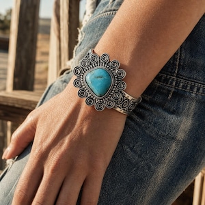 May include: A silver cuff bracelet featuring a central teardrop-shaped turquoise stone surrounded by a decorative border of metal swirls and beadwork. The band of the bracelet displays an intricate engraved pattern along the sides of the metal.