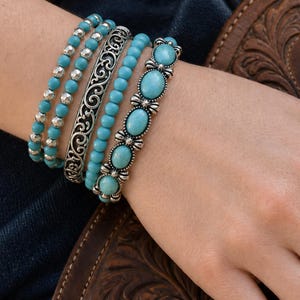May include: A set of four stacked bracelets and a matching ring. The bracelets include two strands of turquoise beads, one silver-toned cuff with intricate scrollwork, and one silver-toned chain featuring oval turquoise cabochons and metal accents.