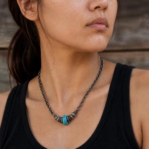Peut inclure: Un collier composé d'une chaîne en argent oxydé foncé avec une série de petits anneaux métalliques texturés entourant une perle unique, lisse et de couleur turquoise vif au centre. La chaîne présente des maillons robustes.