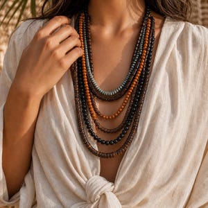 May include: A multi-strand necklace featuring several layers of cylindrical and spherical beads in shades of dark brown, light brown, black, and muted blue. The necklace consists of multiple distinct strands of varying lengths that drape around the neck.