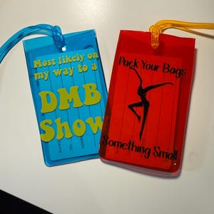 May include: Two rectangular luggage tags. One is blue with yellow text that reads "Most likely on my way to a DMB Show." The other is red with black text that reads "Pack Your Bags Something Small."