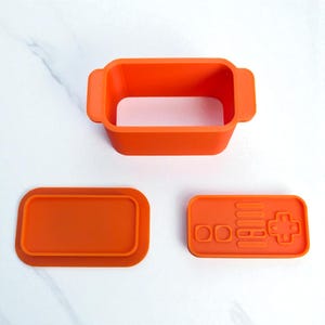 May include: Bright orange silicone baking mold set. Includes a rectangular container with handles, a lid, and a controller-shaped mold. The controller mold features buttons and a directional pad.