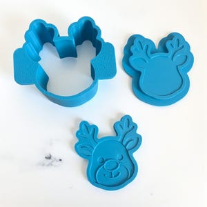May include: Three blue plastic cookie cutters in the shape of a reindeer. One cutter is a complete outline, another is a detailed impression, and the third is a finished reindeer face with antlers, eyes, and a smile. Great for holiday baking.