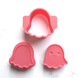 Op de afbeelding: Drie roze plastic koekjessnijders in de vorm van geesten. Eén snijder is een complete geestvorm, een andere is een geestomtrek, en de derde is een geest met een lachend gezicht. De snijders liggen op een wit oppervlak.