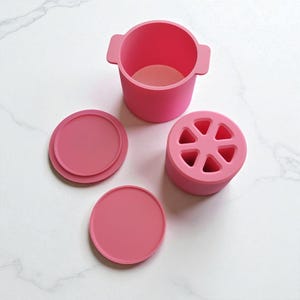 May include: A set of pink silicone kitchen tools, including a small pot with handles, two round lids, and a cylindrical container with six holes. The items are arranged on a white marble surface.