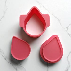 May include: Three pink teardrop-shaped cookie cutters. One cutter has a rectangular top, and the other two are solid teardrop shapes. The cutters are made of plastic and are on a white marble surface.