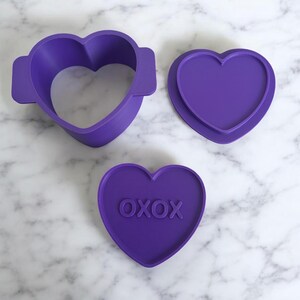 May include: Three purple heart-shaped cookie cutters. One is a frame, another is a solid heart, and the third has the text "OXOX" embossed on it. The cutters are made of plastic and are arranged on a marble surface.