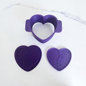 May include: Three purple heart-shaped cookie cutters. One is a solid heart, another has a stitched design, and the third is a heart outline with tabs. These are likely used for baking or crafting projects.