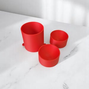 May include: Three red cylindrical containers of varying sizes are arranged on a white surface. The largest container is tall and has a small base. The other two are shorter, with one being wider than the other. The containers are all the same shade of red.