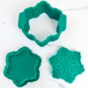 May include: Three teal silicone snowflake-shaped cookie cutters. One is a complete snowflake shape, one is a snowflake outline, and the third is a snowflake with a detailed design. The cutters are on a white marble surface.