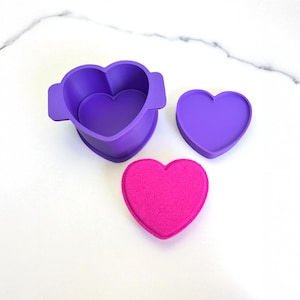 May include: A purple silicone heart-shaped mold with a matching lid and a bright pink heart-shaped item. The mold has handles on the sides. The items are arranged on a white marble surface.