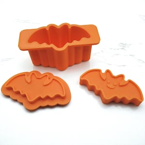 May include: Orange silicone Halloween-themed baking molds. The set includes a rectangular mold shaped like a bat, and two bat-shaped cookie cutters. The molds are made of a flexible, food-safe material.