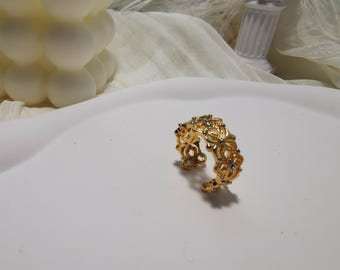 May include: A gold-colored ring with an openwork floral design. The ring features small, clear gemstones and is set on a white surface. The ring is a piece of jewelry.