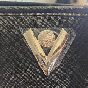 May include: Close-up of a black textured handbag with a gold-colored metal emblem. The emblem is a V-shape with a circular center that has the words "ORLANDI VALENTINO" around a central "V" logo.