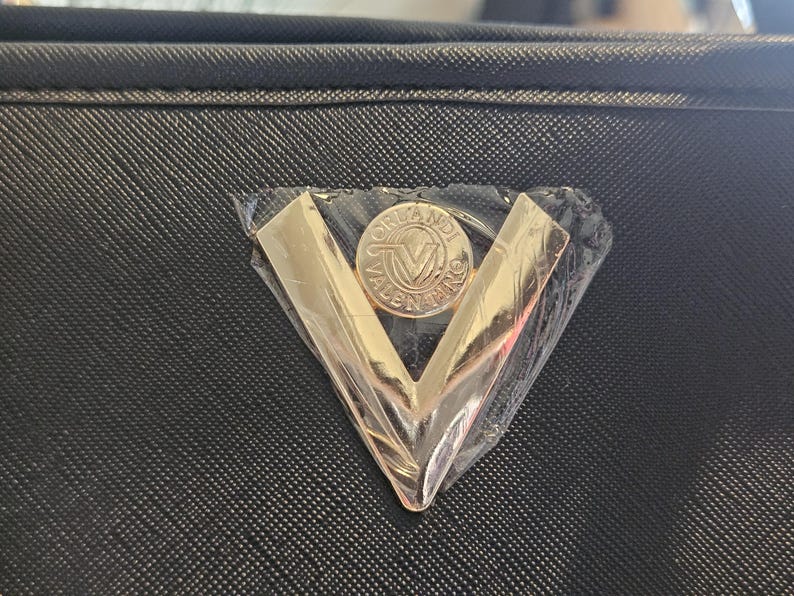 May include: Close-up of a black textured handbag with a gold-colored metal emblem. The emblem is a V-shape with a circular center that has the words "ORLANDI VALENTINO" around a central "V" logo.