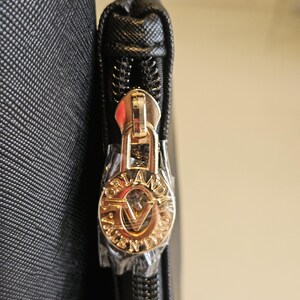 May include: Close-up of a black handbag zipper with a gold-colored pull. The pull features the brand name "ORLANDO VALENTINO" in a circular design. The zipper is partially open, revealing the interior.