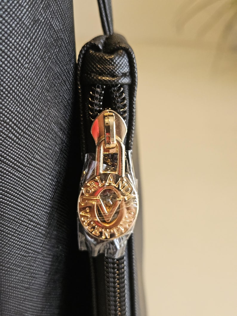May include: Close-up of a black handbag zipper with a gold-colored pull. The pull features the brand name "ORLANDO VALENTINO" in a circular design. The zipper is partially open, revealing the interior.