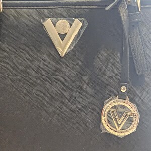 May include: Black tote bag with gold-colored hardware, including a triangular emblem and a circular charm with the letter "V". The bag has a textured surface and a strap.