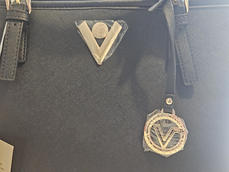 May include: Black tote bag with gold-colored hardware, including a triangular emblem and a circular charm with the letter "V". The bag has a textured surface and a strap.