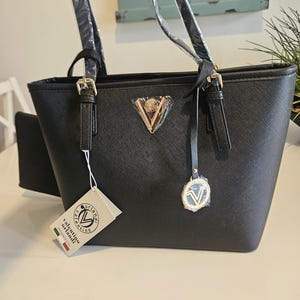May include: Black tote bag with gold-colored hardware, including buckles and a triangular emblem. The bag has two handles and a small attached pouch. A tag reads "valentino orlandi".