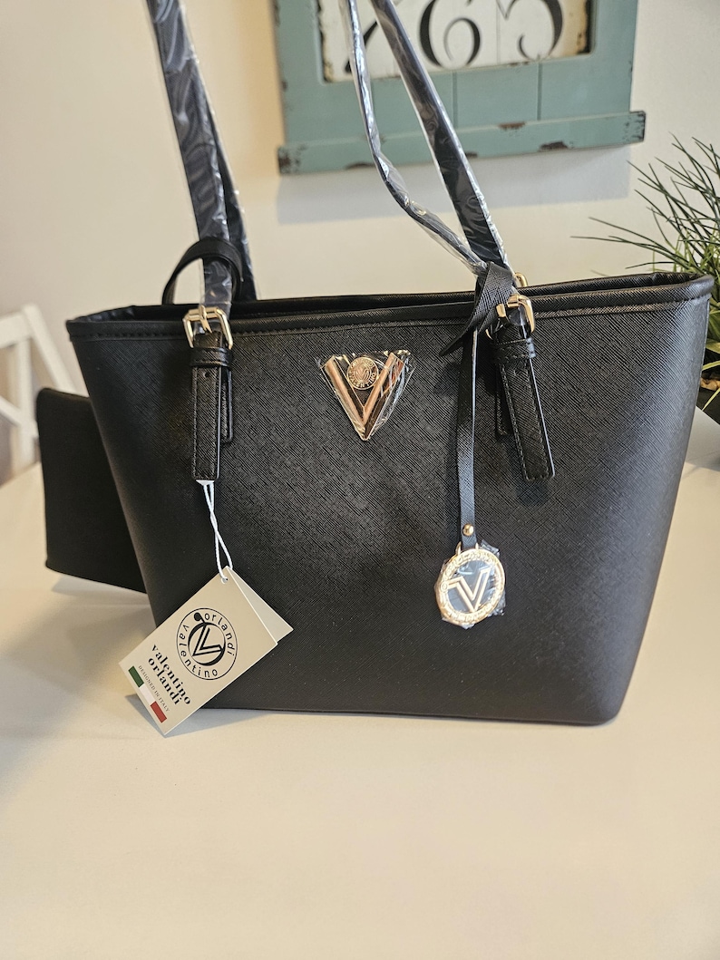 May include: Black tote bag with gold-colored hardware, including buckles and a triangular emblem. The bag has two handles and a small attached pouch. A tag reads "valentino orlandi".