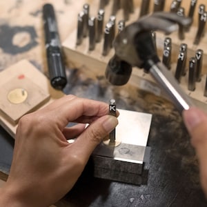 May include: A close-up of a person using a hammer to stamp the letter "K" onto a small, round, gold-colored metal disc. The disc rests on a metal block. Other metal letter stamps and a black marker are in the background.