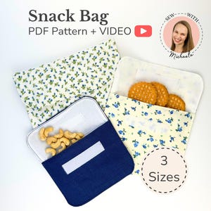 May include: Three fabric snack bags in different patterns and colors, including a solid navy blue, a white floral print, and a yellow floral print. The bags feature white hook-and-loop closures and contain various snacks like cashews and crackers.