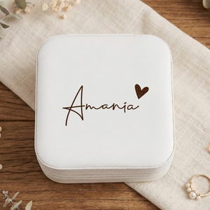 May include: A white square jewellery box with the name "Amania" in a handwritten font and a small brown heart. The box is on a white cloth with gold jewellery and dried flowers in the background.