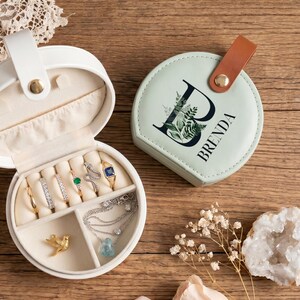 May include: Two round jewelry cases, one white and open with rings, a gold bird charm, and a silver necklace inside, and one sage green closed case featuring a brown leather tab and the letter B with floral details printed on the lid.