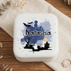 May include: A square white jewelry box featuring a Halloween-themed graphic with the name Natasha printed in black. The design includes blue watercolor splashes, black silhouettes of bats, a tree, a haunted house, tombstones, and white ghosts.