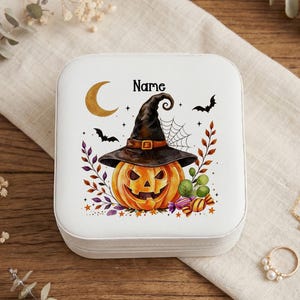 May include: A square white jewelry box featuring a printed illustration of a jack-o'-lantern wearing a black witch hat, a crescent moon, bats, a spider web, and colorful autumn foliage. The word Name is printed in black above the pumpkin.