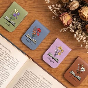 May include: Four leather bookmarks with floral designs and names: Kallie, Paulina, Brayan, and Johanna. Each bookmark features an open book illustration. The bookmarks are arranged on a wooden surface near an open book and dried flowers.