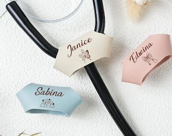 Può includere: Uno stetoscopio con tre targhette personalizzate. Le targhette sono beige, rosa e blu, ognuna con un nome e una piccola grafica. La targhetta beige ha il nome "Janice" e una farfalla. La targhetta rosa ha il nome "Edwina" e una grafica di stetoscopio. La targhetta blu ha il nome "Sabina" e un arcobaleno.