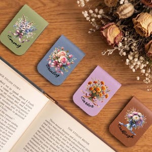 Puede incluir: Cuatro marcadores de cuero personalizados con diseños florales y nombres: Emmy, Brinley, Carter y Oakleigh. Los marcadores son en tonos de verde, azul, lavanda y marrón. Un libro y flores secas también están en la imagen.
